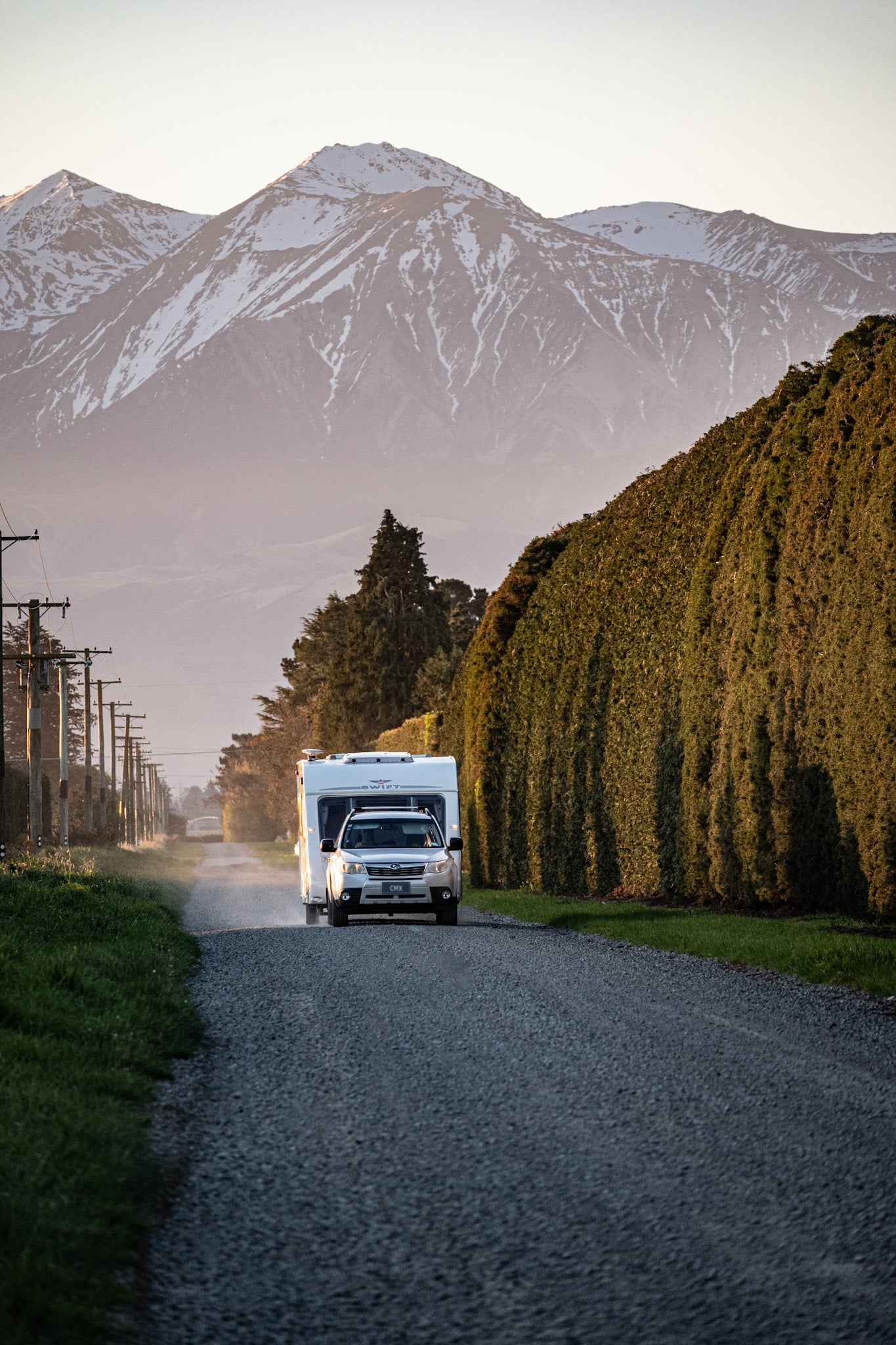 Car towing a caravan along a gravel road towards snow-capped mountains